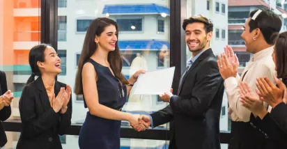 Image shows a coworker presenting an award to another coworker in front of a group of teammates.